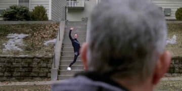 Escena de "El peor vecino del mundo" con Tom Hanks observando a su vecina Marisol, interpretada por Mariana Treviño, quien le saluda con entusiasmo desde unas escaleras frente a su casa.