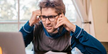 Hombre joven serio, con gafas, telefoneando frente al portátil al enterarse del bloqueo de su cuenta bancaria por falta de documentación