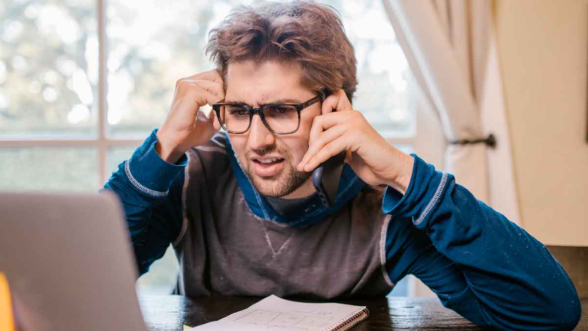 Hombre joven serio, con gafas, telefoneando frente al portátil al enterarse del bloqueo de su cuenta bancaria por falta de documentación