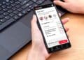 Persona sosteniendo un smartphone con la app Cuenta Digital de la Comunidad de Madrid abierta, gestionando trámites públicos junto a un ordenador portátil