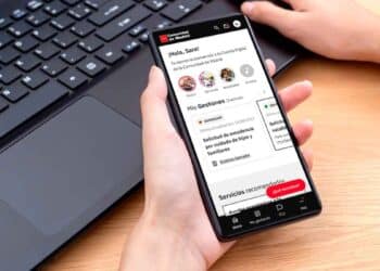 Persona sosteniendo un smartphone con la app Cuenta Digital de la Comunidad de Madrid abierta, gestionando trámites públicos junto a un ordenador portátil