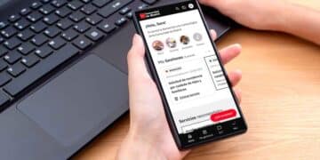 Persona sosteniendo un smartphone con la app Cuenta Digital de la Comunidad de Madrid abierta, gestionando trámites públicos junto a un ordenador portátil
