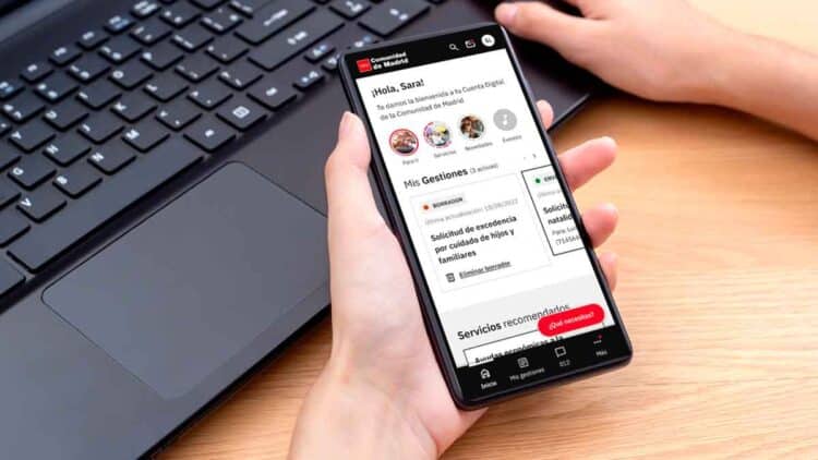 Persona sosteniendo un smartphone con la app Cuenta Digital de la Comunidad de Madrid abierta, gestionando trámites públicos junto a un ordenador portátil