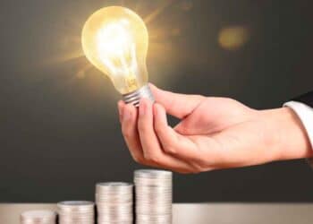 Mano sujetando una bombilla encendida junto a pilas de monedas que simbolizan el ahorro en la factura de la luz gracias al Bono Social Eléctrico