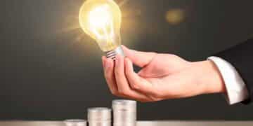 Mano sujetando una bombilla encendida junto a pilas de monedas que simbolizan el ahorro en la factura de la luz gracias al Bono Social Eléctrico
