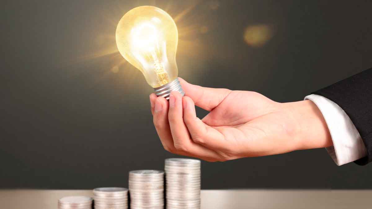 Mano sujetando una bombilla encendida junto a pilas de monedas que simbolizan el ahorro en la factura de la luz gracias al Bono Social Eléctrico