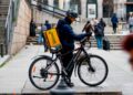 Repartidor de Glovo en bicicleta con mochila amarilla, esperando pedido en una calle céntrica de España en 2025