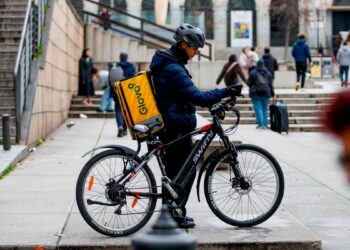 Repartidor de Glovo en bicicleta con mochila amarilla, esperando pedido en una calle céntrica de España en 2025