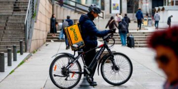 Repartidor de Glovo en bicicleta con mochila amarilla, esperando pedido en una calle céntrica de España en 2025