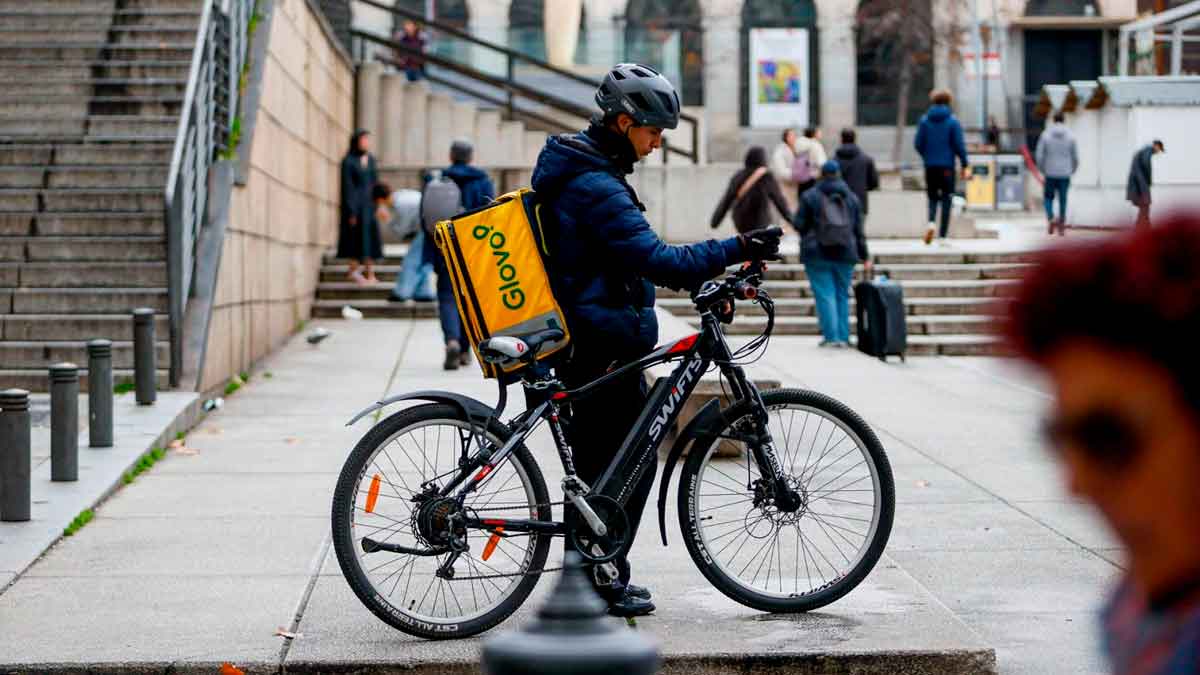 Repartidor de Glovo en bicicleta con mochila amarilla, esperando pedido en una calle céntrica de España en 2025