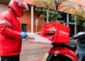Repartidor de Telepizza con chaqueta y casco rojos guardando cajas de pizza en el baúl de su moto en plena calle