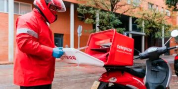 Repartidor de Telepizza con chaqueta y casco rojos guardando cajas de pizza en el baúl de su moto en plena calle