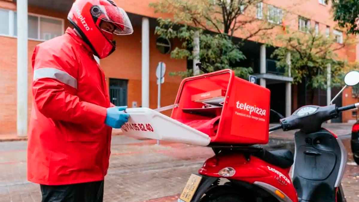 Repartidor de Telepizza con chaqueta y casco rojos guardando cajas de pizza en el baúl de su moto en plena calle