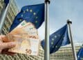 Mano mostrando billetes de euro frente a banderas de la UE, símbolo de las ayudas europeas que España pierde por burocracia.