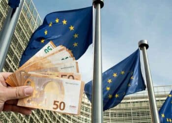 Mano mostrando billetes de euro frente a banderas de la UE, símbolo de las ayudas europeas que España pierde por burocracia.