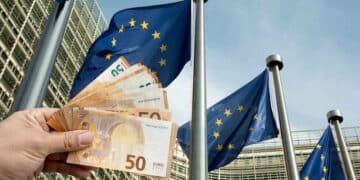 Mano mostrando billetes de euro frente a banderas de la UE, símbolo de las ayudas europeas que España pierde por burocracia.