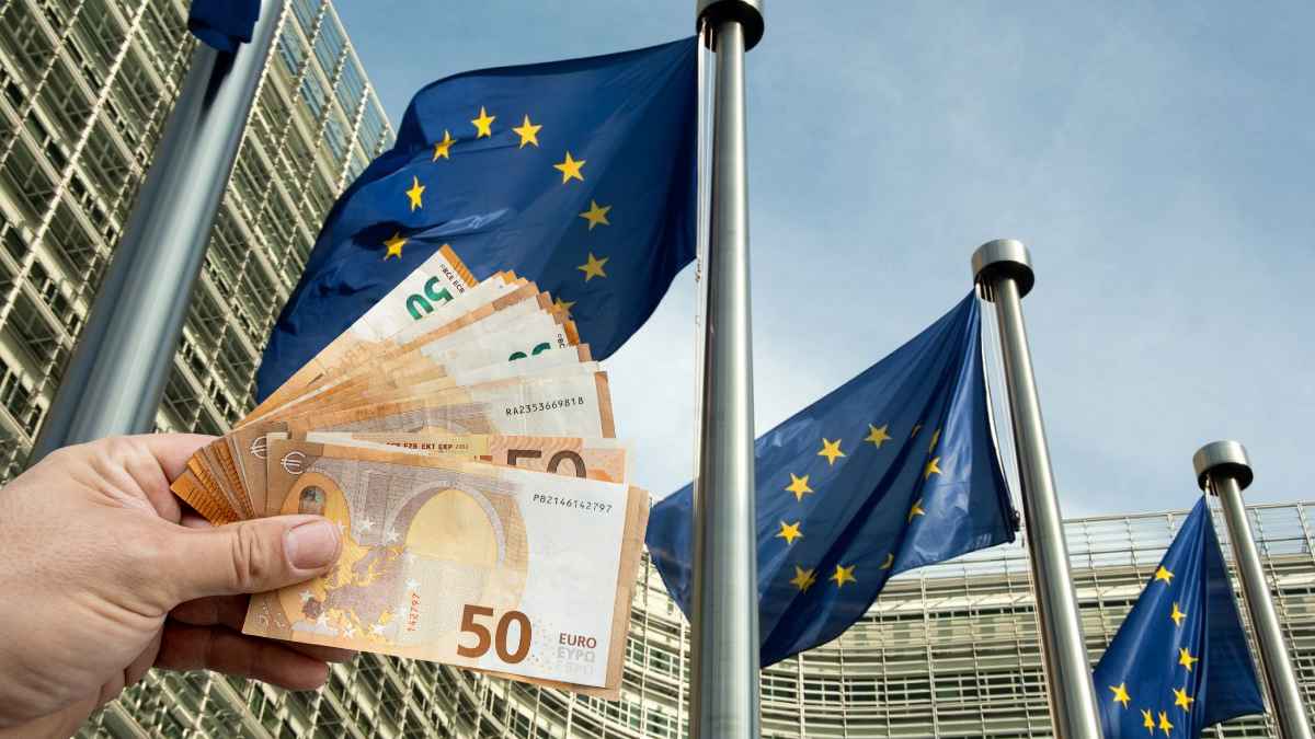Mano mostrando billetes de euro frente a banderas de la UE, símbolo de las ayudas europeas que España pierde por burocracia.