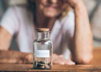 Tarro de cristal con monedas y la palabra “pensión” delante de una mujer mayor pensativa, símbolo del ahorro para jubilarse en 2026 con 15 años cotizados