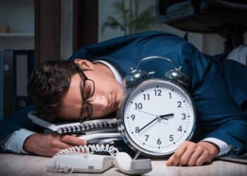 Empleado agotado dormido junto a un gran reloj, símbolo de las horas extra obligatorias según el Estatuto de los Trabajadores.