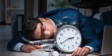 Empleado agotado dormido junto a un gran reloj, símbolo de las horas extra obligatorias según el Estatuto de los Trabajadores.