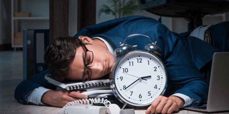 Empleado agotado dormido junto a un gran reloj, símbolo de las horas extra obligatorias según el Estatuto de los Trabajadores.
