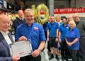 Empleado Kevin Parsons sostiene un certificado de 40 años de servicio, rodeado de compañeros y globos dorados “40” en el supermercado Alliance