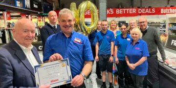 Empleado Kevin Parsons sostiene un certificado de 40 años de servicio, rodeado de compañeros y globos dorados “40” en el supermercado Alliance