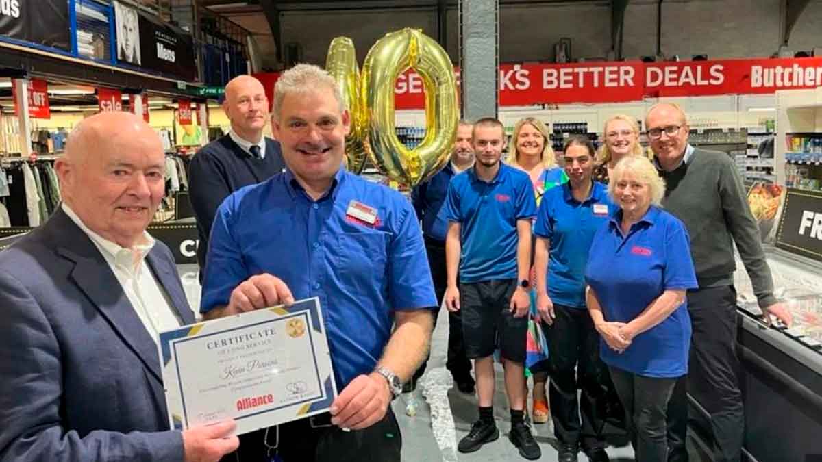 Empleado Kevin Parsons sostiene un certificado de 40 años de servicio, rodeado de compañeros y globos dorados “40” en el supermercado Alliance