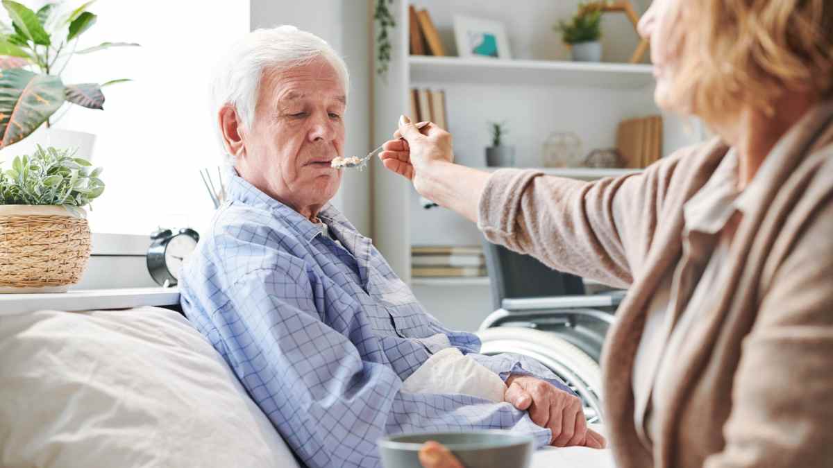 Cuidadora alimenta a un hombre mayor dependiente en casa; allegados, amigos y vecinos podrán recibir ayudas económicas por su cuidado.