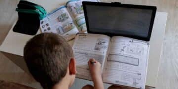 Niño haciendo deberes con tablet y libro, ejemplo de deberes digitales en colegios.