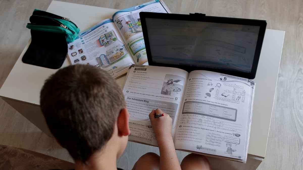 Niño haciendo deberes con tablet y libro, ejemplo de deberes digitales en colegios.