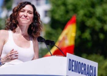 Isabel Díaz Ayuso en acto público en Madrid, donde se ve a una mujer haciendo un discurso sobre democracia con bandera de España