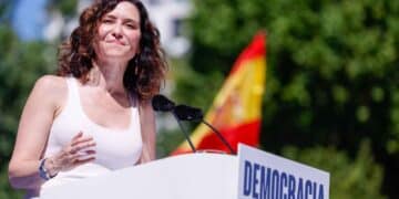 Isabel Díaz Ayuso en acto público en Madrid, donde se ve a una mujer haciendo un discurso sobre democracia con bandera de España