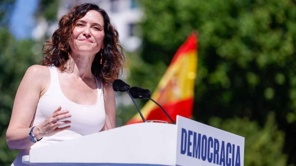 Isabel Díaz Ayuso en acto público en Madrid, donde se ve a una mujer haciendo un discurso sobre democracia con bandera de España