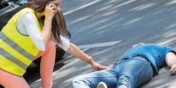 Mujer con chaleco reflectante llama al 112 y socorre a un herido tendido en la carretera tras un accidente de tráfico, aplicando la regla PAS.