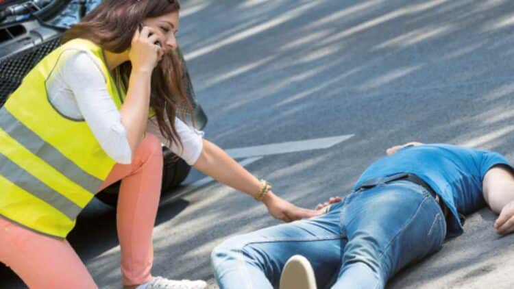 Mujer con chaleco reflectante llama al 112 y socorre a un herido tendido en la carretera tras un accidente de tráfico, aplicando la regla PAS.