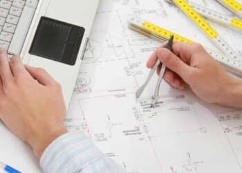 Manos de estudiante trabajando en planos de arquitectura con ordenador portátil y compás.
