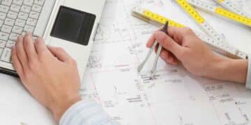 Manos de estudiante trabajando en planos de arquitectura con ordenador portátil y compás.