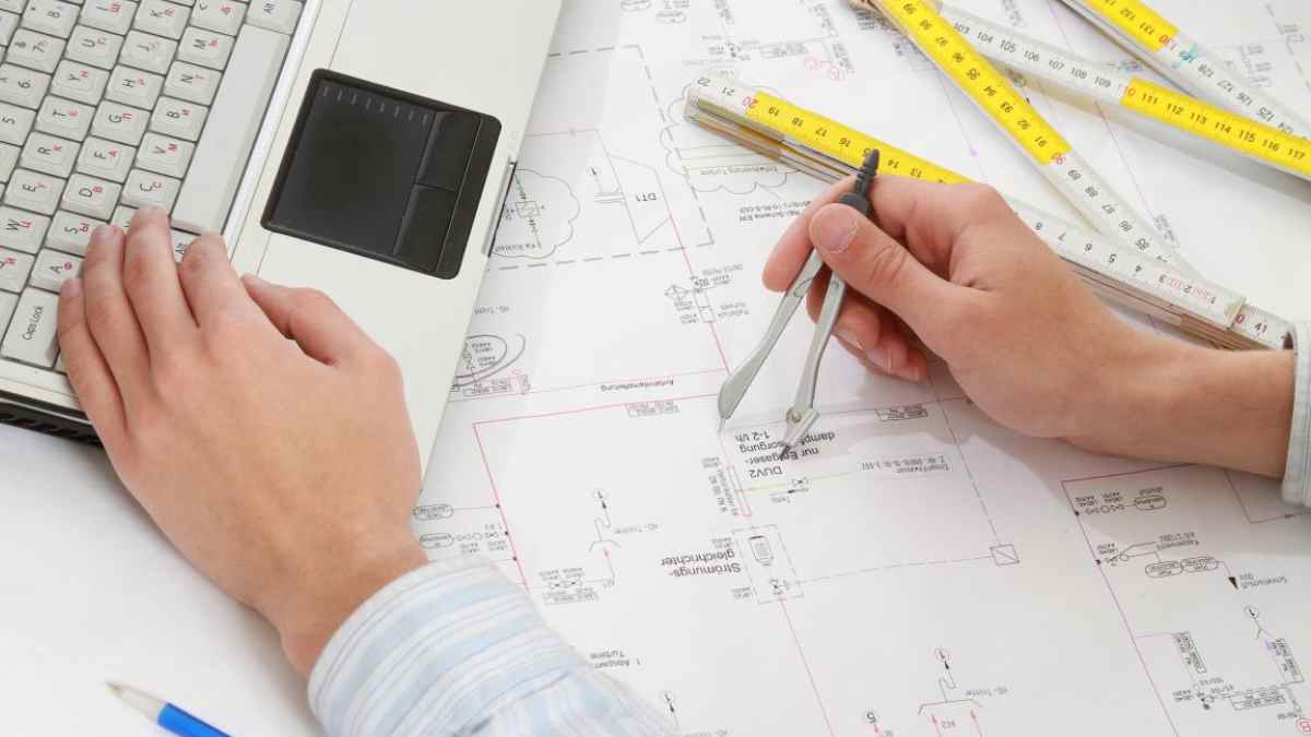 Manos de estudiante trabajando en planos de arquitectura con ordenador portátil y compás.