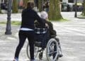 Mujer empujando silla de ruedas a persona mayor en parque.