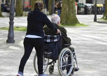 Mujer empujando silla de ruedas a persona mayor en parque.