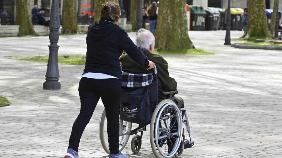 Mujer empujando silla de ruedas a persona mayor en parque.