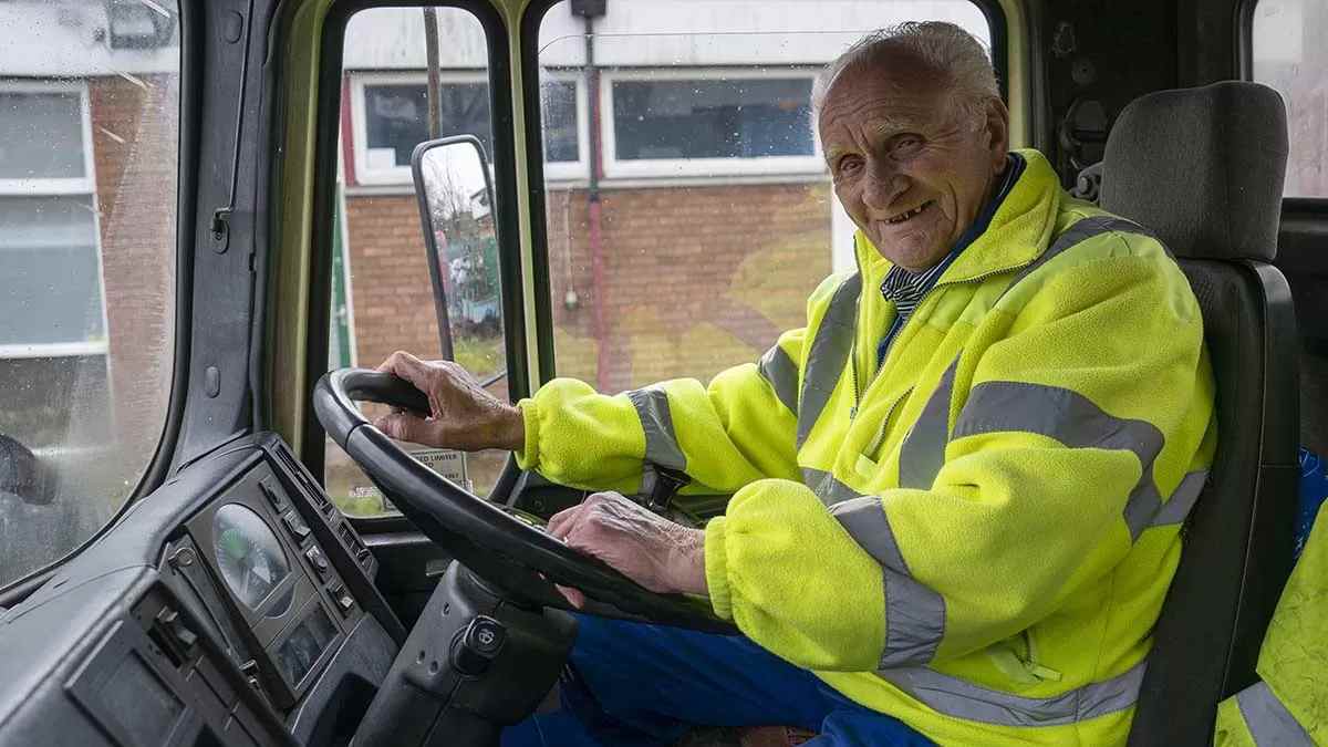 Funcionario mayor conduciendo camión con chaleco reflectante antes de jubilarse a los 72 años.