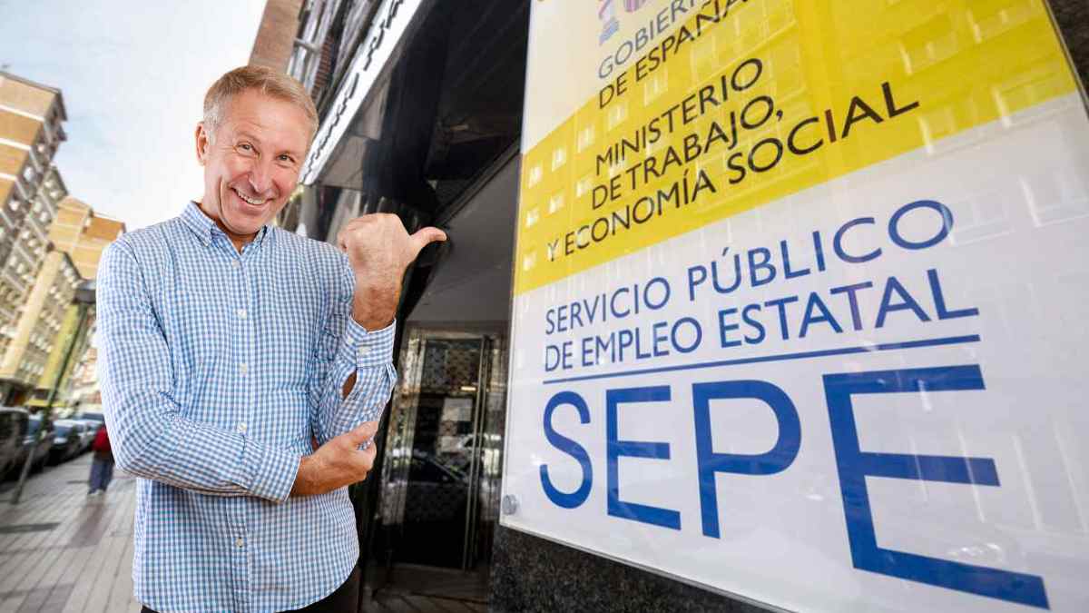 Hombre mayor señalando cartel del SEPE sobre subsidio de 482 euros para mayores de 52 años.