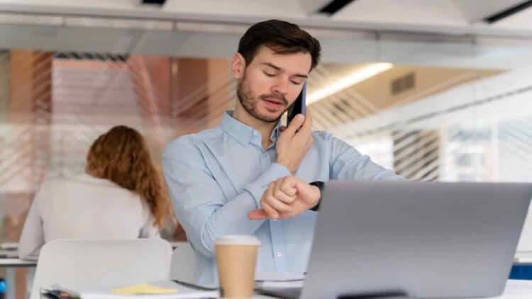 Empleado revisando su horario laboral mientras trabaja en oficina y habla por teléfono.