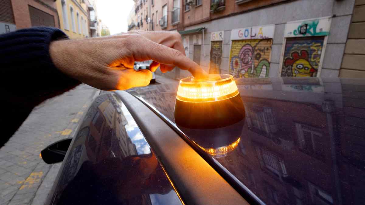 Mano activando una baliza V-16 encendida sobre el techo de un coche en una calle urbana.