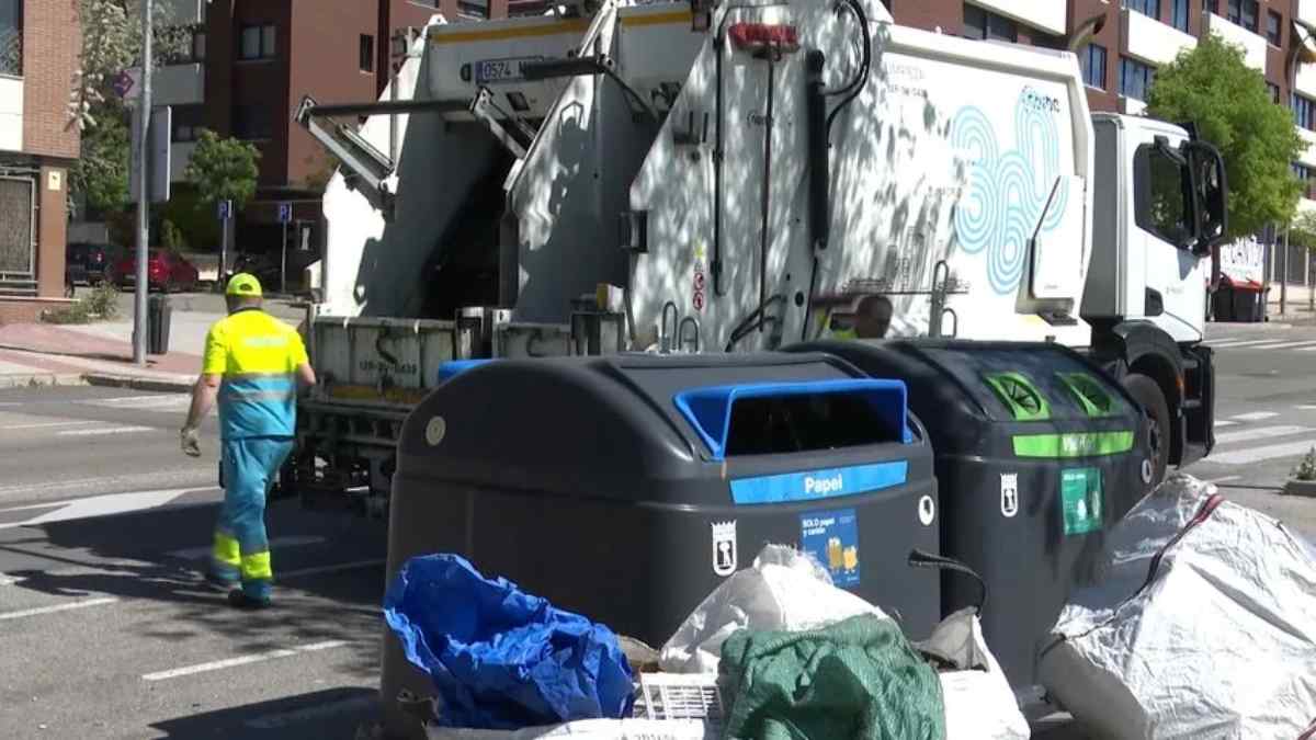 Trabajador de recogida de basura junto a camión y contenedores en la vía pública.
