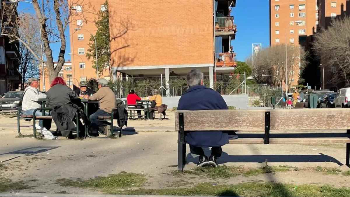 Vecinos sentados en un parque de un barrio de Madrid con edificios residenciales al fondo.