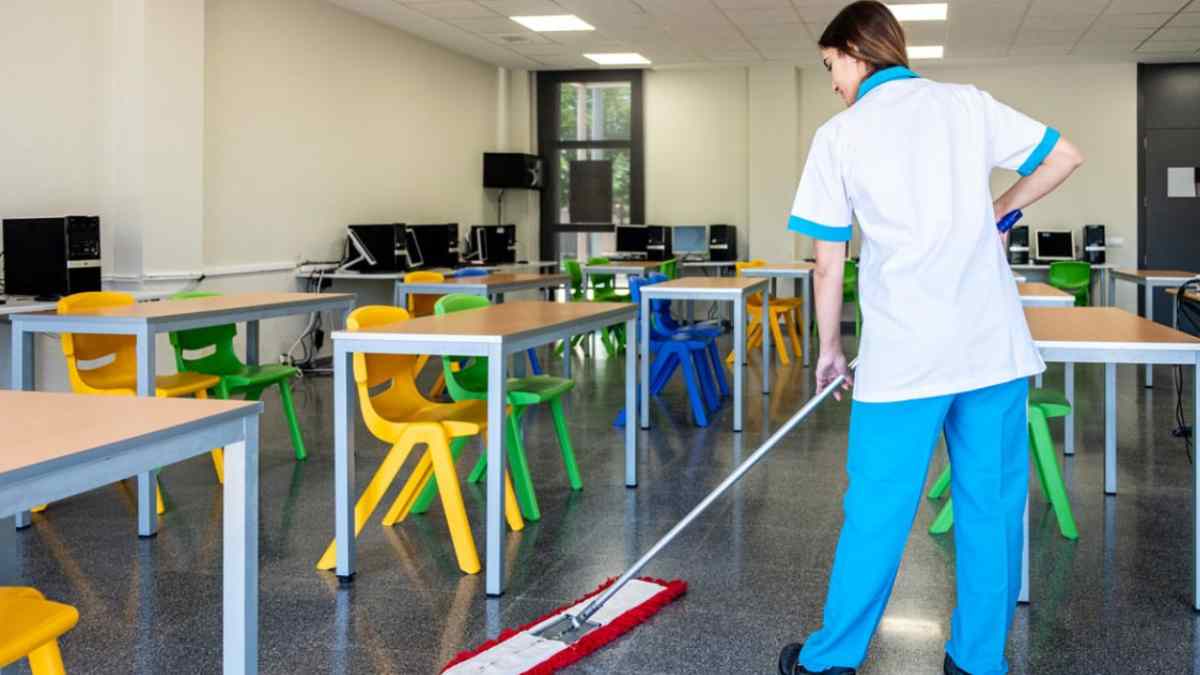 Trabajadora de limpieza realizando tareas en un aula de colegio con mesas y sillas de colores.