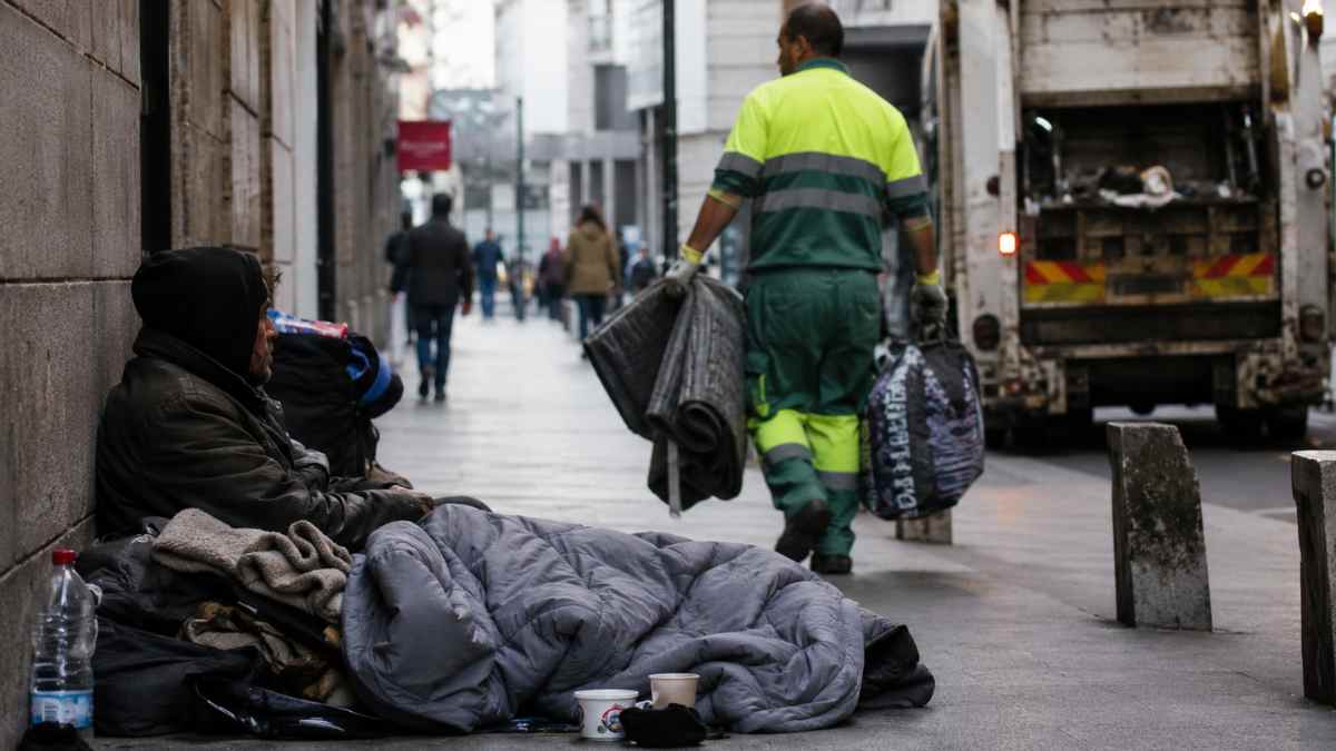 Persona sin hogar junto a sus pertenencias mientras un operario de limpieza las retira en una calle de Madrid